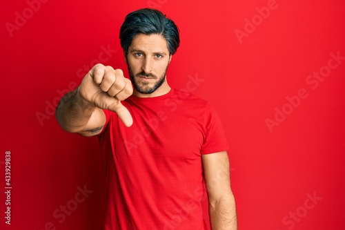 Young hispanic man wearing casual red t shirt looking unhappy and angry showing rejection and negative with thumbs down gesture. bad expression.
