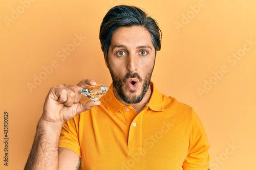 Photography Young hispanic man holding brilliant diamond stone scared and amazed with open m