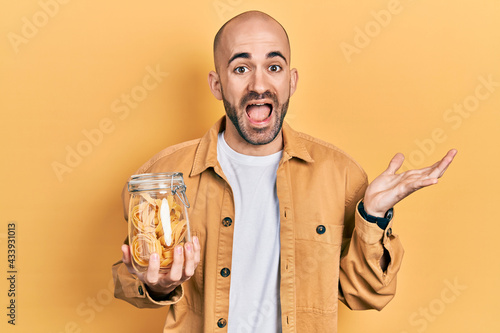 Young bald man holding jar with uncooked pasta celebrating achievement with happy smile and winner expression with raised hand