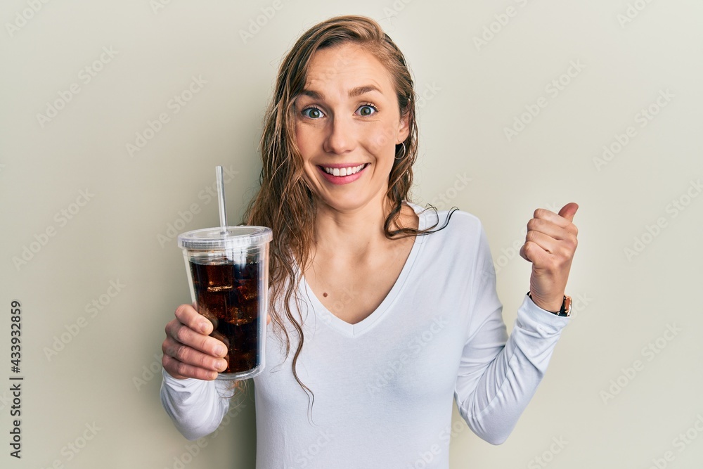 Young blonde woman drinking glass of soda beverage pointing thumb up to the side smiling happy with open mouth Young blonde woman drinking glass of soda beverage pointing thumb up to the side smiling happy with open mouth