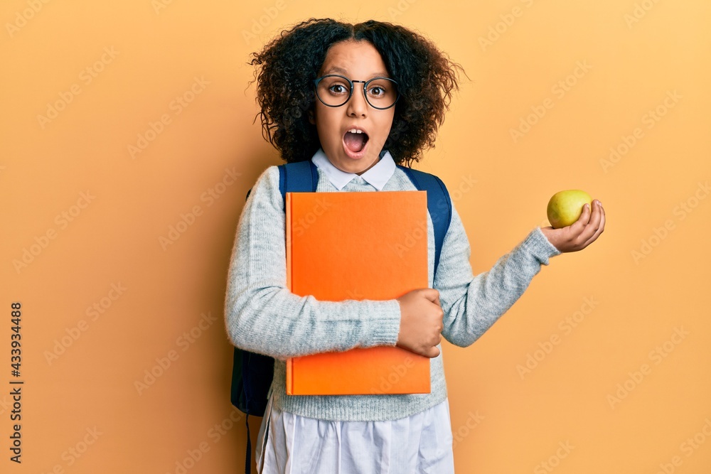 Young little girl with afro hair wearing school bag holding books and green apple celebrating crazy and amazed for success with open eyes screaming excited. Young little girl with afro hair wearing school bag holding books and green apple celebrating crazy and amazed for success with open eyes screaming excited.
