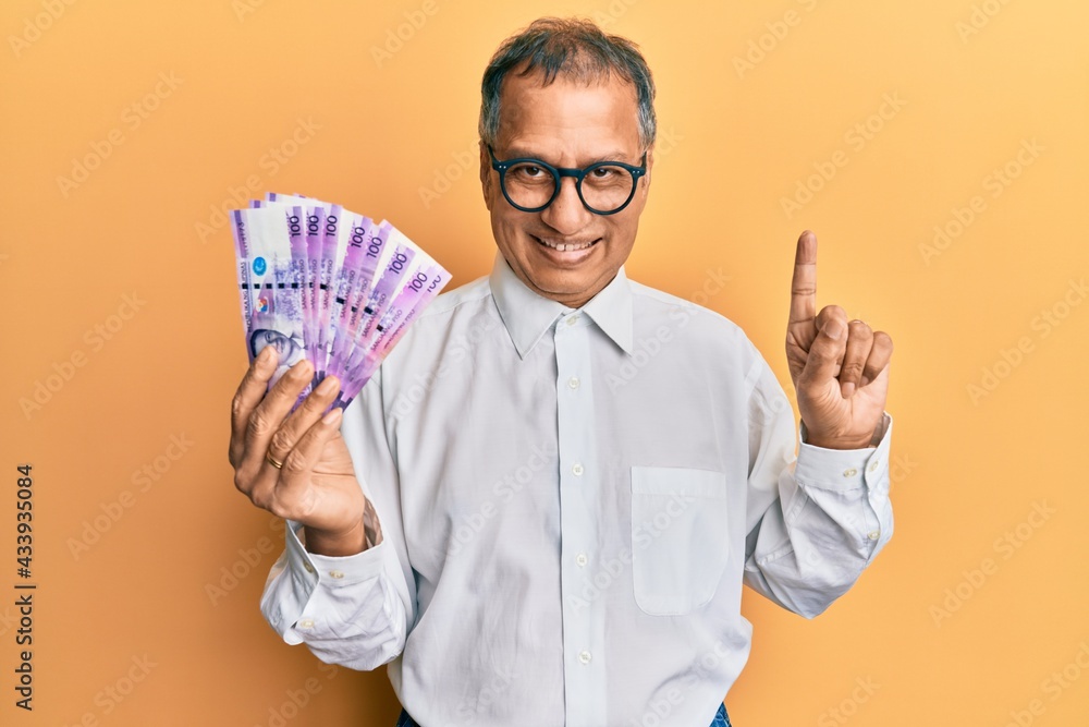 Middle age indian man holding 100 philippine peso banknotes smiling with an idea or question pointing finger with happy face, number one Middle age indian man holding 100 philippine peso banknotes smiling with an idea or question pointing finger with happy face, number one