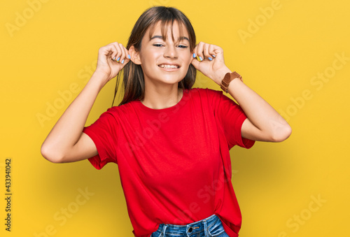 Teenager caucasian girl wearing casual red t shirt smiling pulling ears with fingers, funny gesture. audition problem