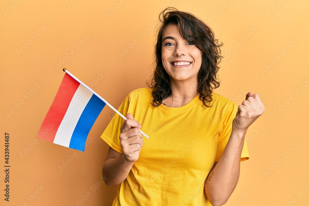Young hispanic woman holding holland flag screaming proud, celebrating victory and success very excited with raised arm Young hispanic woman holding holland flag screaming proud, celebrating victory and success very excited with raised arm