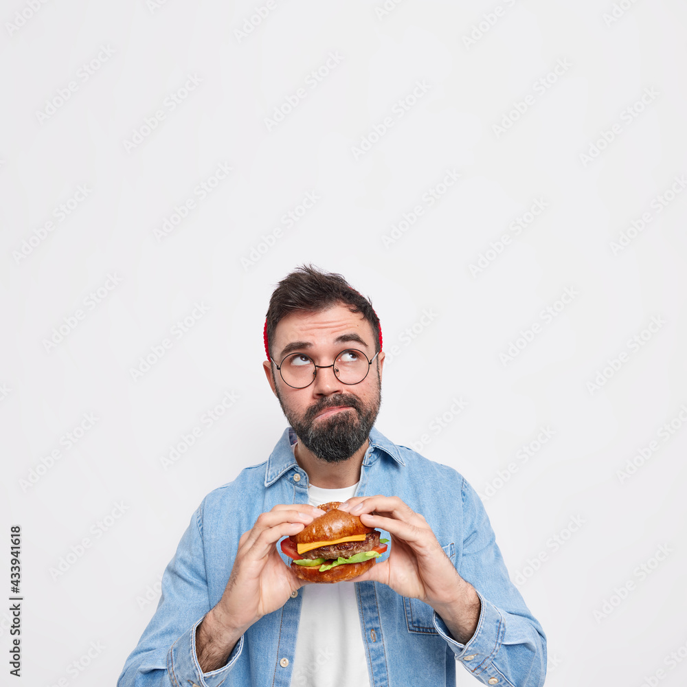 Vertical shot of pensive bearded man holds appetizing hamburger focused above thinks deeply about something wears round spectacles and denim shirt blank copy space overhead for advertisement Vertical shot of pensive bearded man holds appetizing hamburger focused above thinks deeply about something wears round spectacles and denim shirt blank copy space overhead for advertisement
