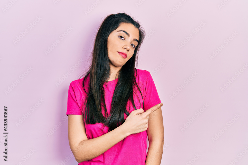 Young hispanic girl wearing casual pink t shirt pointing with hand finger to the side showing advertisement, serious and calm face Young hispanic girl wearing casual pink t shirt pointing with hand finger to the side showing advertisement, serious and calm face