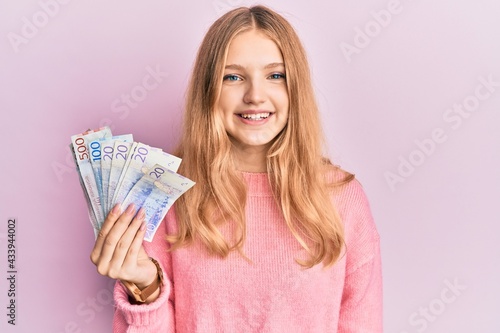 Beautiful young caucasian girl holding swedish krona banknotes looking positive and happy standing and smiling with a confident smile showing teeth