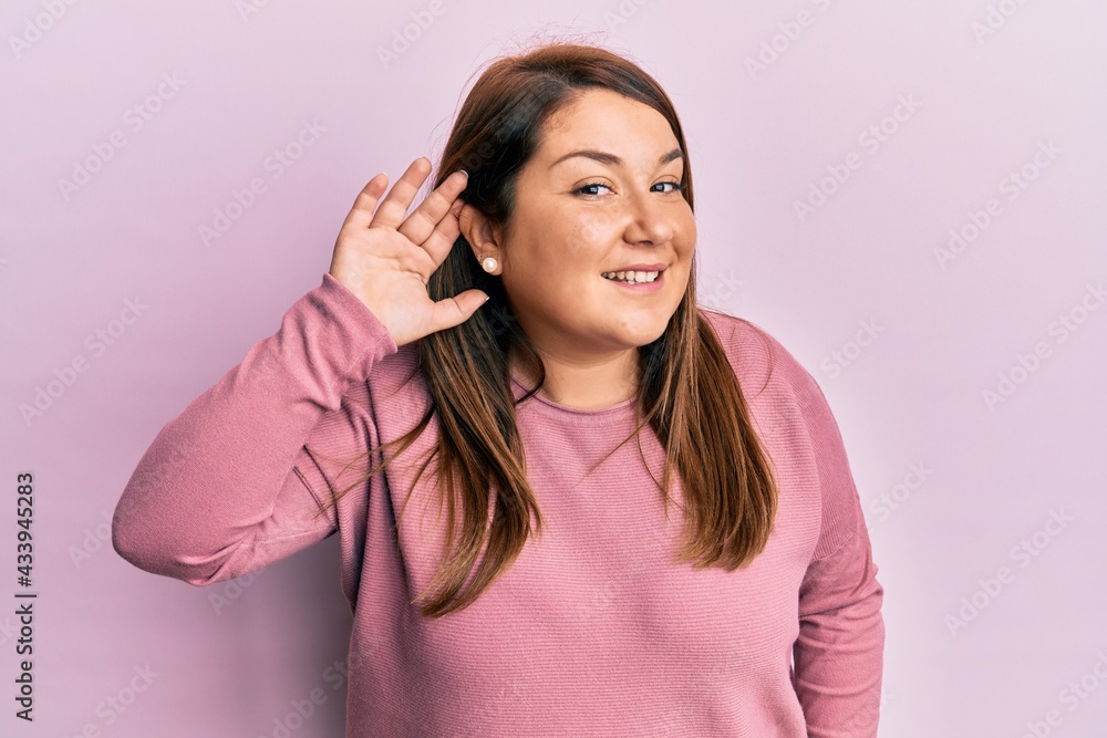 Beautiful brunette plus size woman wearing casual winter sweater smiling with hand over ear listening and hearing to rumor or gossip. deafness concept. Beautiful brunette plus size woman wearing casual winter sweater smiling with hand over ear listening and hearing to rumor or gossip. deafness concept.