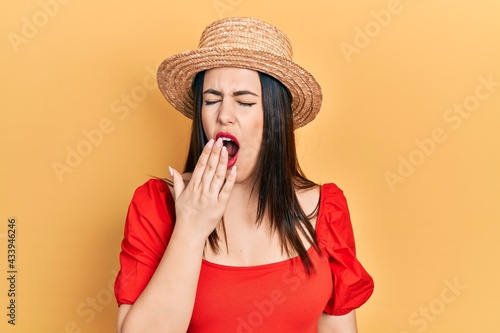 Young hispanic woman wearing summer hat bored yawning tired covering mouth with hand. restless and sleepiness.