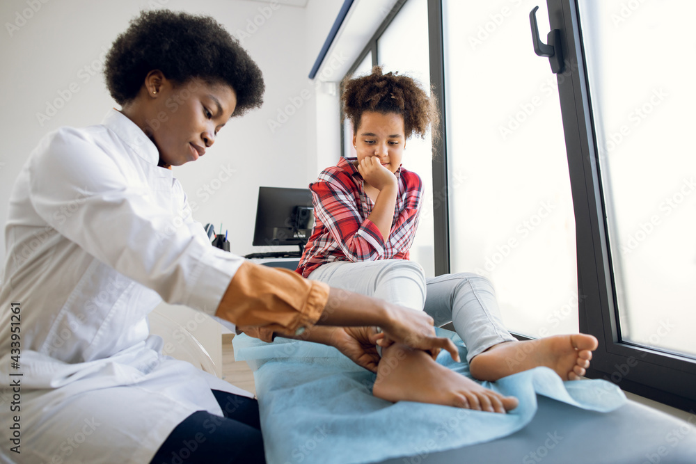 Cute curly mixed race teen 12-aged girl, sitting on the couch at modern clinic, while young 30-aged female African doctor orthopedist rheumatologist examining patient's leg Cute curly mixed race teen 12-aged girl, sitting on the couch at modern clinic, while young 30-aged female African doctor orthopedist rheumatologist examining patient's leg