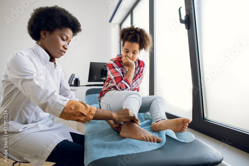 Cute curly mixed race teen 12-aged girl, sitting on the couch at modern clinic, while young 30-aged female African doctor orthopedist rheumatologist examining patient's leg