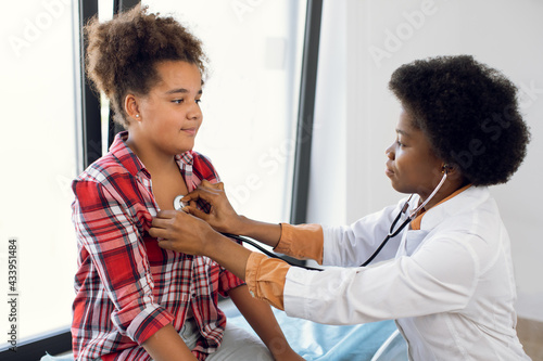 African female pediatrician with stethoscope auscultating cute teen mixed race girl patient. Black physician, checks heartbeat and lungs breathing of kid, making checkup before vaccination photo