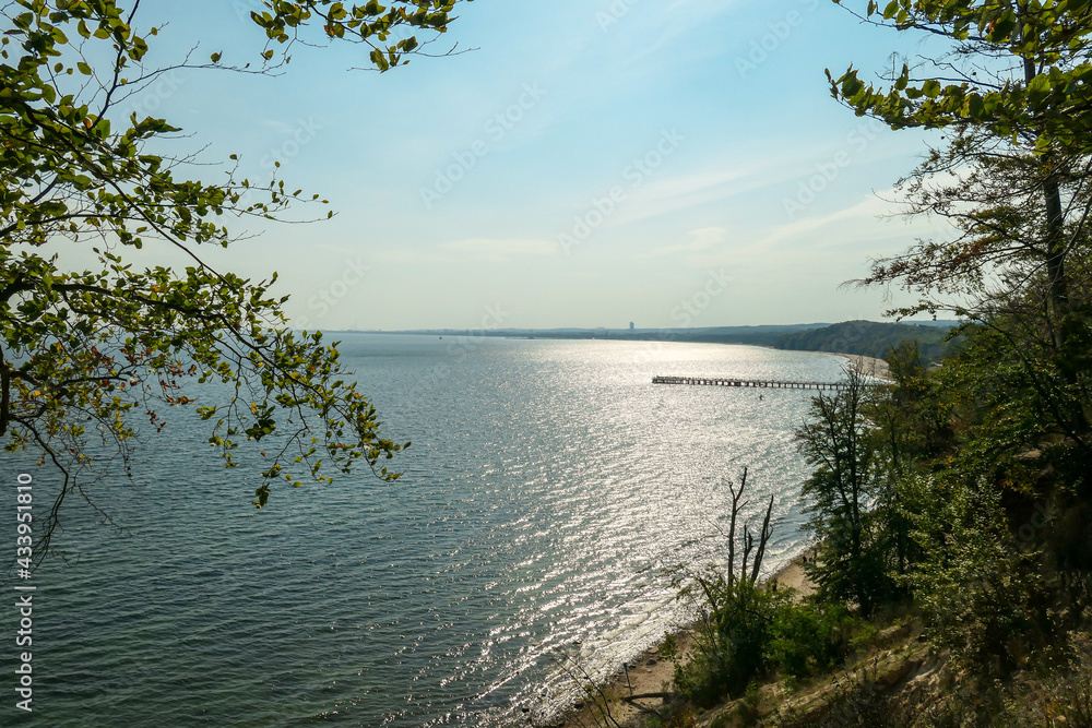 A panoramic view on the costal line in Gdynia, Poland, seen from a small cliff above the sea level.  There is a white pier going into the calm Baltic Sea. High grass overgrowing the shore. Idyllic