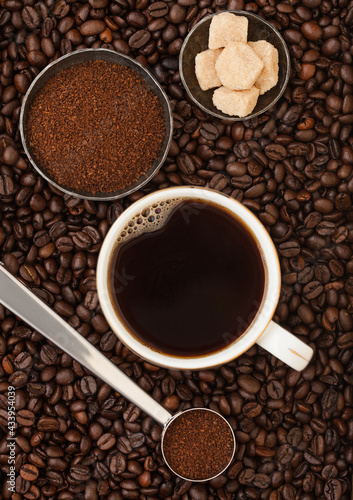 Coffee cup with ground coffee in steel scoop and cane sugar inside fresh coffee beans background.