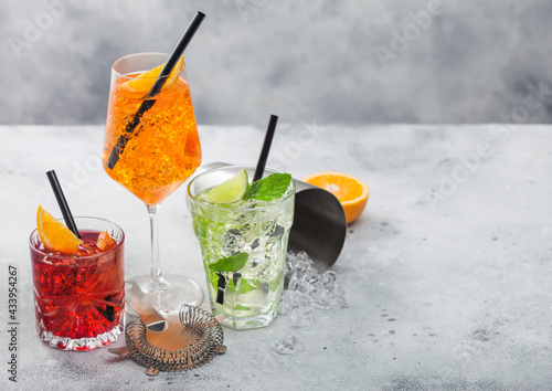 Glasses of spritz,mojito and negroni cocktails with ice cubes and lime and orange slices with mint leaf and black straw on light background with strainer and shaker.