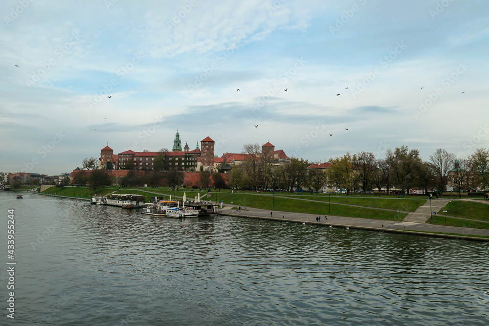 Obraz premium A panoramic view on the iconic Wawel Castle in Cracow, Poland and the Vistula river flowing under the castle. A few birds flying above the river. A few boats docked on the rivers bend. City tour.