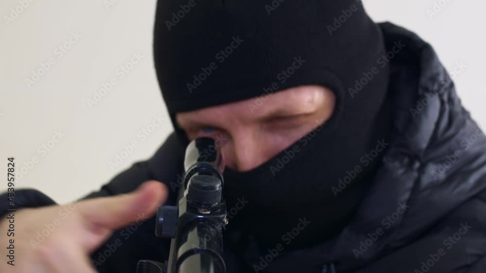 The assassin in the black blaclava raises the sniper rifle, adjusts the telescopic sight and looks for the target. Turns an evil gaze into the camera. Contract murder. Close-up. The assassin in the black blaclava raises the sniper rifle, adjusts the telescopic sight and looks for the target. Turns an evil gaze into the camera. Contract murder. Close-up.
