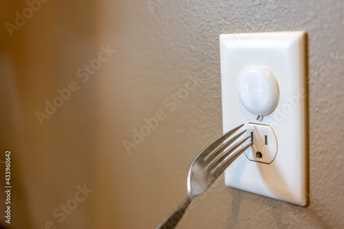 Selective focus on edge of a metal fork about to be inserted into an open electrical outlet socket. The top outlet has a plastic insert for childproofing safety protection. photo