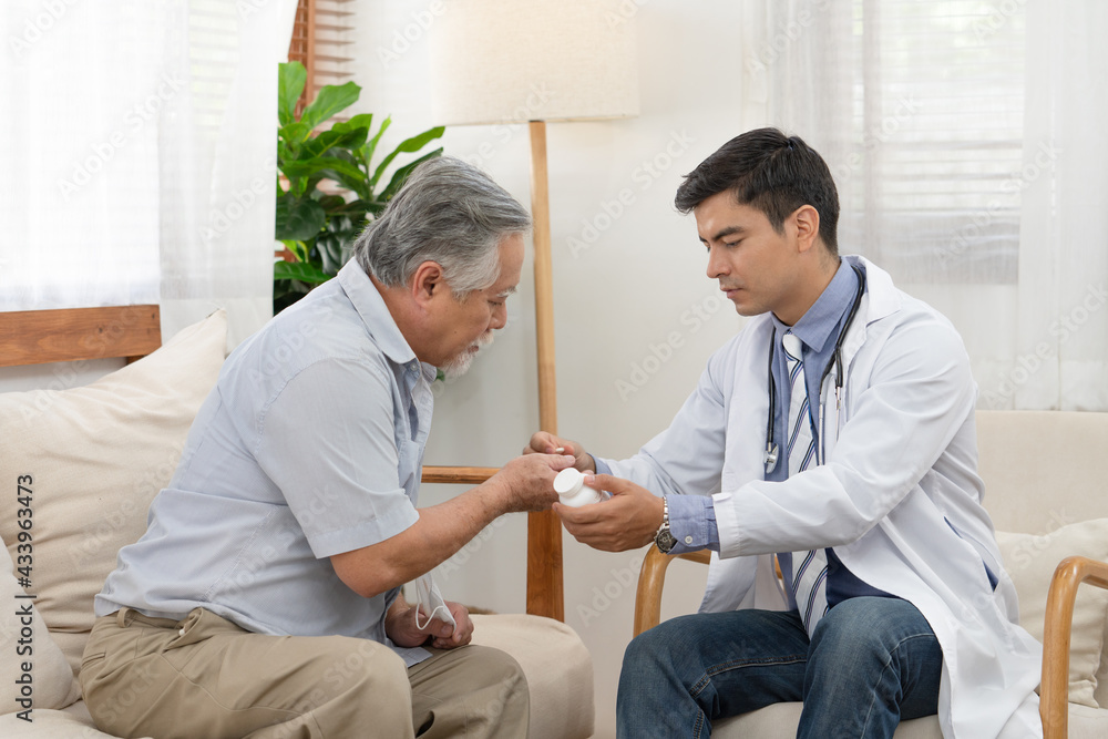 Obraz premium Doctor hold bottle of pills and explaining using medication to Asian elderly man patient sitting on sofa in living room after medical examination. Doctor provided the service to visit at home.