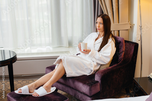 A young beautiful girl in a white coat drinks coffee in her hotel room.