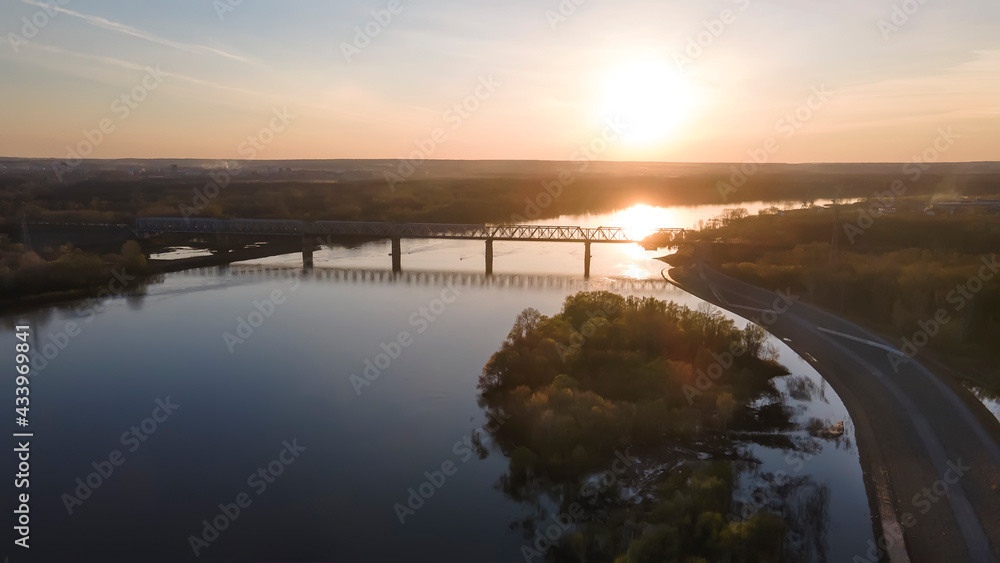 A beautiful bright sun sets at sunset. Bridge over a large river. Beautiful evening nature. Water and an island in the river. Sunny evening colorful landscape.