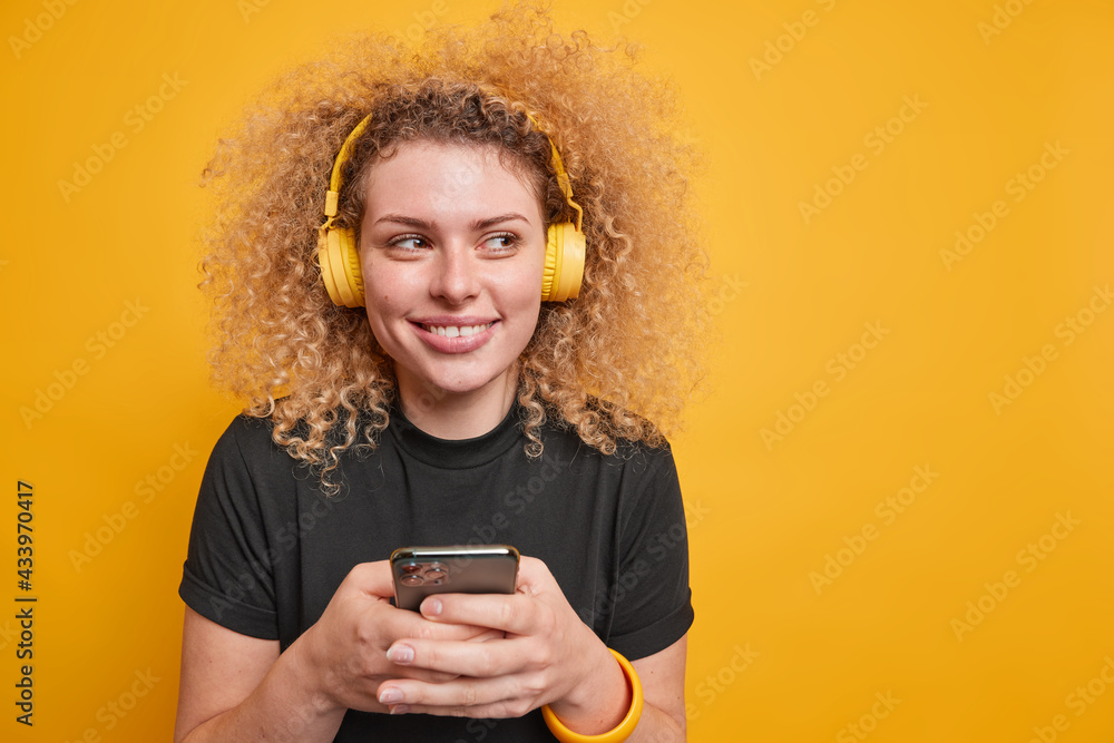 Pretty smiling curly haired young woman looks away happily listens music uses smartphone enjoys good sound quality wears casual black t shirt isolated over yellow background with blank space Pretty smiling curly haired young woman looks away happily listens music uses smartphone enjoys good sound quality wears casual black t shirt isolated over yellow background with blank space