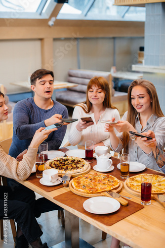 A group of young cheerful friends is sitting in a cafe talking and taking selfies on the phone. Lunch at the pizzeria.