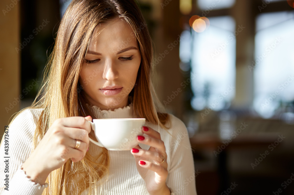 Beautiful attractive business woman in a white blouse drinks cofee in the cafe. Coffee break. Morning light falls from the window Beautiful attractive business woman in a white blouse drinks cofee in the cafe. Coffee break. Morning light falls from the window