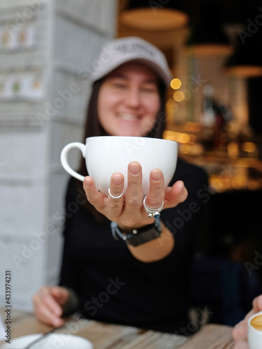 A girl sits in a coffee shop and holds a white coffee cup on her outstretched hand. Smile. The face is in the background and out of focus.