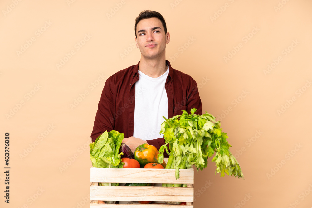 Farmer with freshly picked vegetables in a box isolated on beige background making doubts gesture looking side Farmer with freshly picked vegetables in a box isolated on beige background making doubts gesture looking side