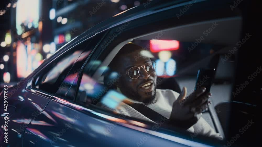 Stylish Black Man in Glasses is Commuting Home in a Backseat of a Taxi at Night. Handsome Male Making a Video Call on a Smartphone while in Transfer Car on City Street with Working Neon Signs. Stylish Black Man in Glasses is Commuting Home in a Backseat of a Taxi at Night. Handsome Male Making a Video Call on a Smartphone while in Transfer Car on City Street with Working Neon Signs.