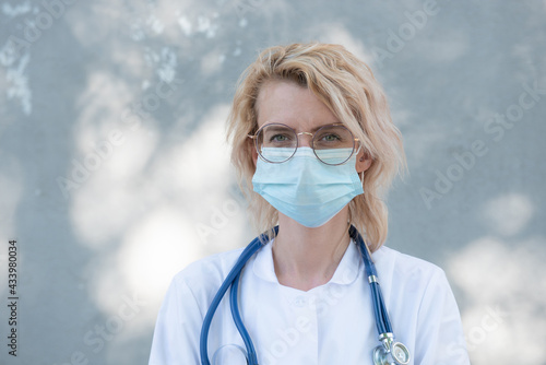 young beautiful girl doctor in a white coat with a phonendoscope in glasses on a gray background in a medical mask smiling
