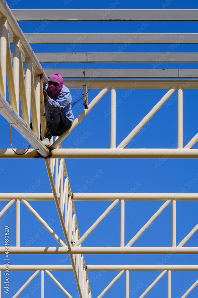 Welder is welding metal on roof structure of warehouse building in construction site against blue sky in vertical frame