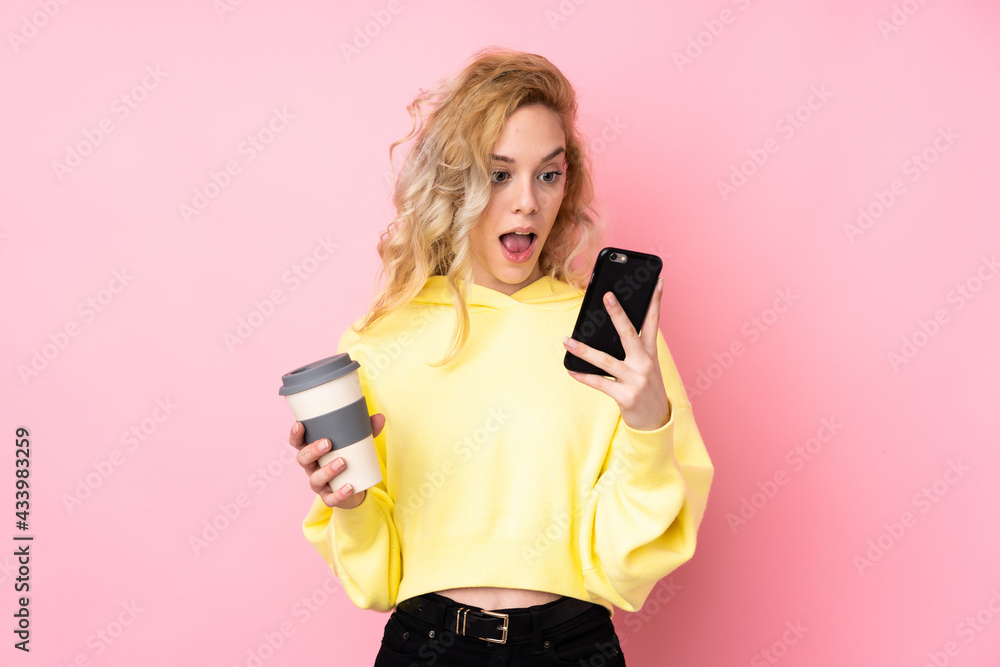 Young blonde woman wearing a sweatshirt isolated on pink background holding coffee to take away and a mobile Young blonde woman wearing a sweatshirt isolated on pink background holding coffee to take away and a mobile