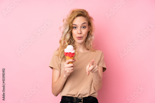 Young blonde woman holding a cornet ice cream isolated on pink background surprised and pointing front