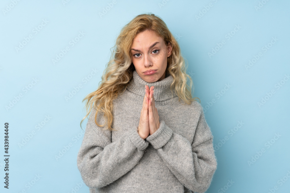 Young blonde woman wearing a sweater isolated on blue background keeps palm together. Person asks for something Young blonde woman wearing a sweater isolated on blue background keeps palm together. Person asks for something