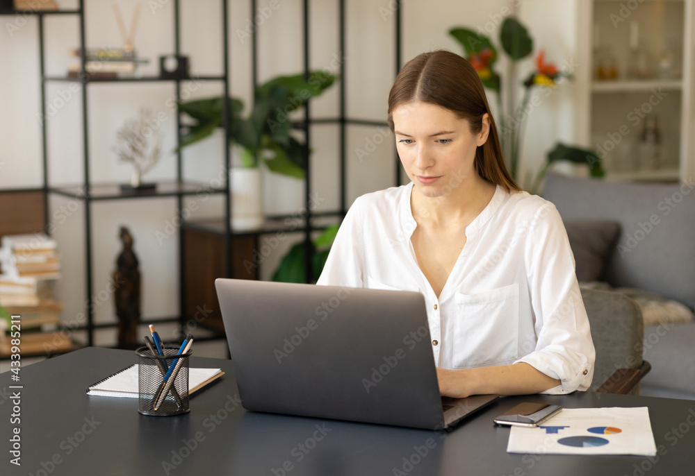 Millennial girl sit at desk at home or office working or studying on laptop in living room Millennial girl sit at desk at home or office working or studying on laptop in living room