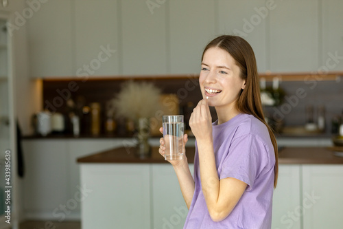 Fotografia Young beautiful smiling caucasian woman holding vitamin pill and glass of water standing in the kitchen at home