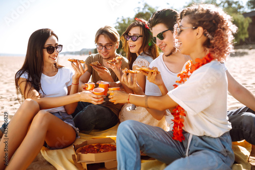 Group of young friends having picnic  eating pizza  toasting with beerus at the beach. Happy friends resting together sitting near the sea. Fast food concept. Beach holiday and summer vacation concept