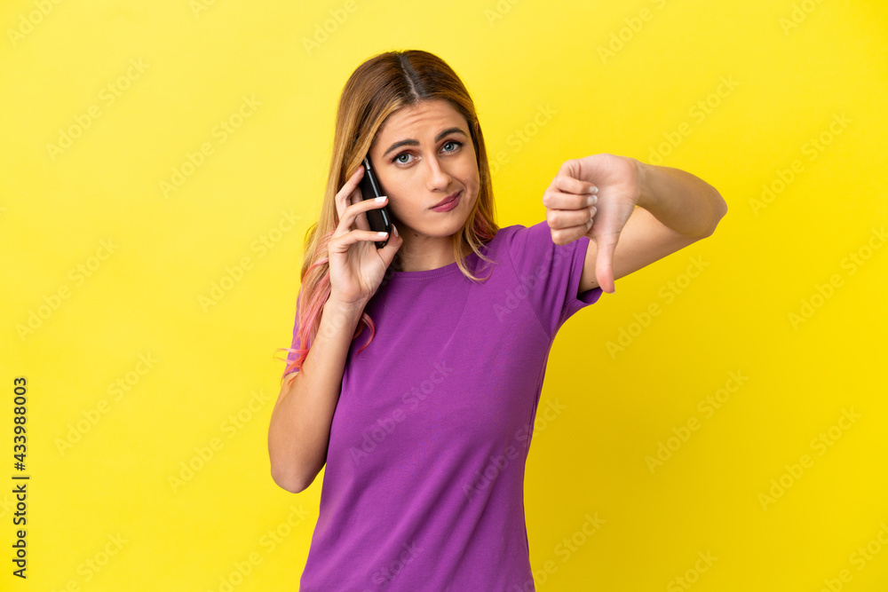 Young woman using mobile phone over isolated yellow background showing thumb down with negative expression Young woman using mobile phone over isolated yellow background showing thumb down with negative expression