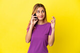 Young woman using mobile phone over isolated yellow background with fingers crossing and wishing the best