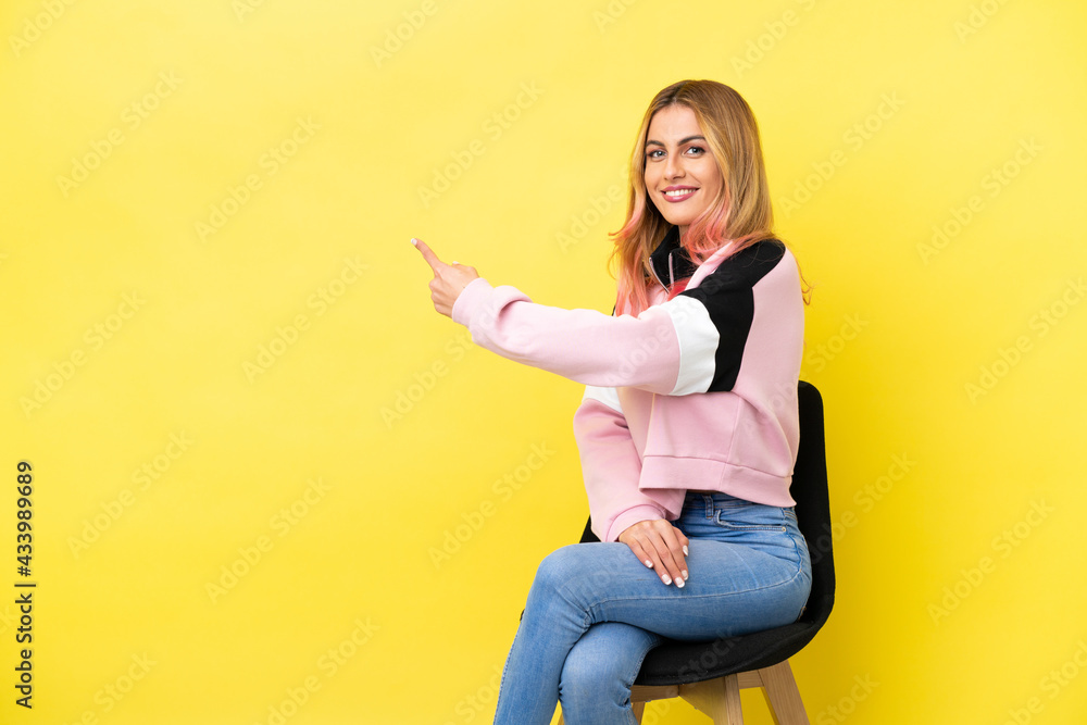 Young woman sitting on a chair over isolated yellow background pointing back Young woman sitting on a chair over isolated yellow background pointing back