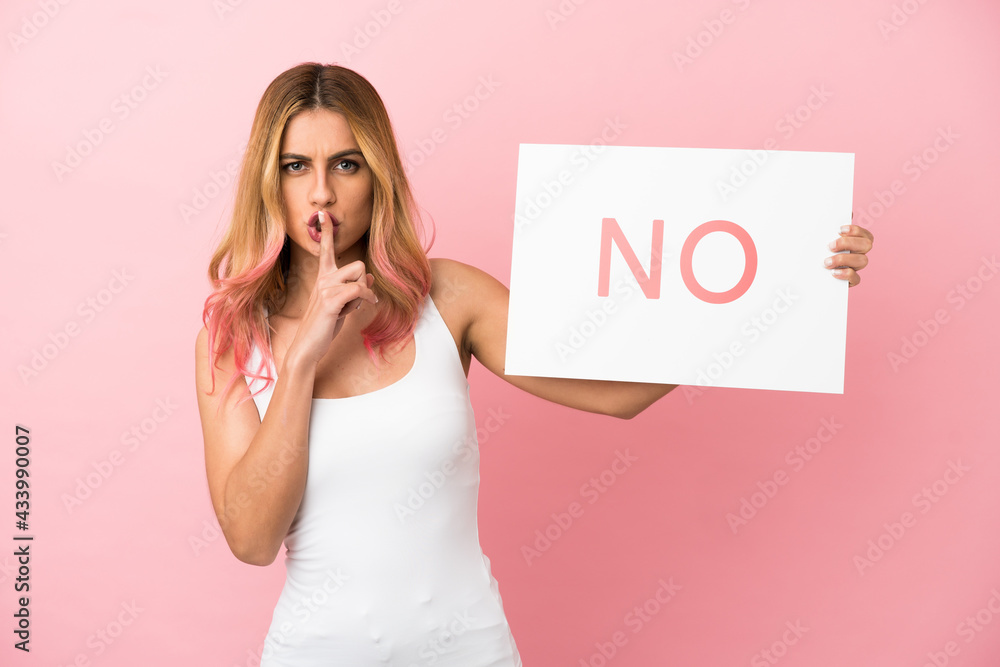 Young woman over isolated pink background holding a placard with text NO doing silence gesture Young woman over isolated pink background holding a placard with text NO doing silence gesture