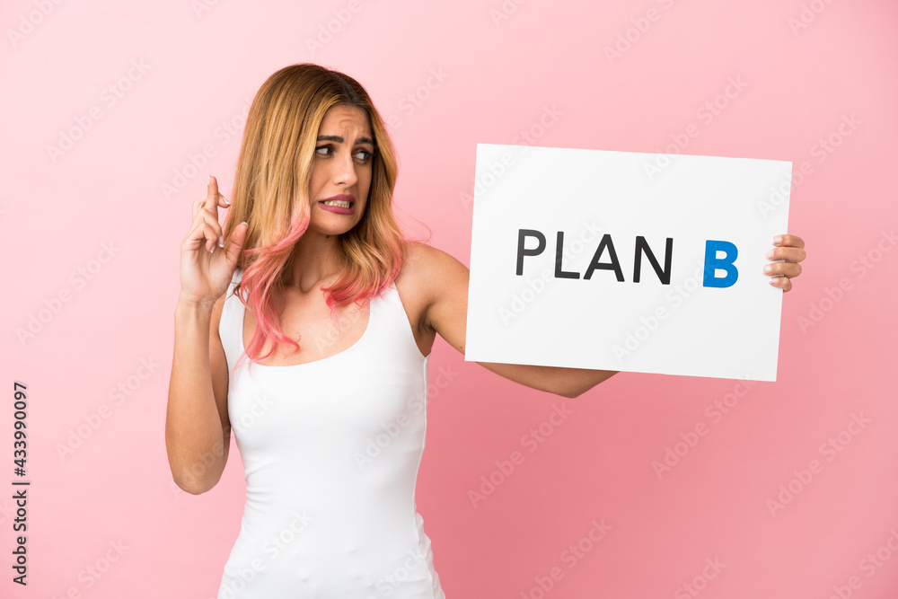Young woman over isolated pink background holding a placard with the message PLAN B with fingers crossing Young woman over isolated pink background holding a placard with the message PLAN B with fingers crossing