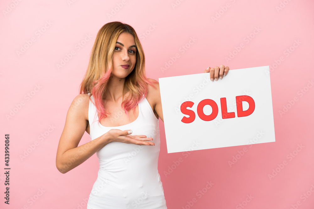Young woman over isolated pink background holding a placard with text SOLD and pointing it Young woman over isolated pink background holding a placard with text SOLD and pointing it