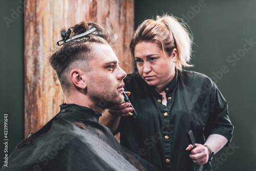 Girl hairdresser makes a haircut to a young guy using a hair clipper in a barbershop