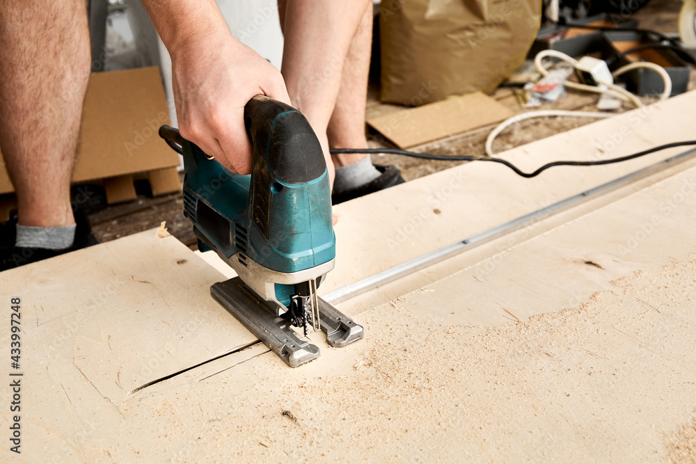 Male hands of construction worker sawing and cutting parts with an electric jigsaw from plywood. The cut process. DIY building of interior decoration with fretsaw. Rental tool. Copy space. Equipment