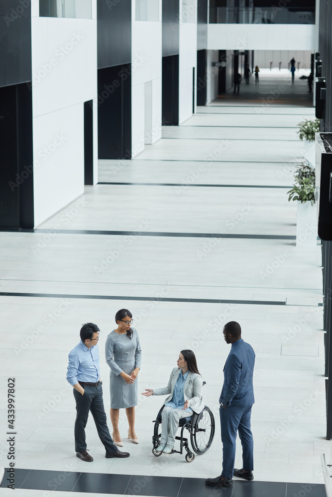 Fototapeta premium Vertical wide angle view at young businesswoman in wheelchair talking to diverse group of people in office lobby, copy space