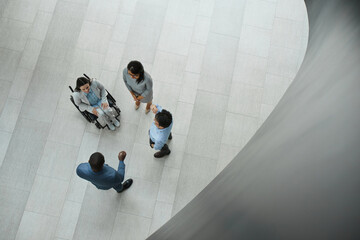 Graphic top down view at young businesswoman in wheelchair talking to diverse group of people in office lobby, copy space