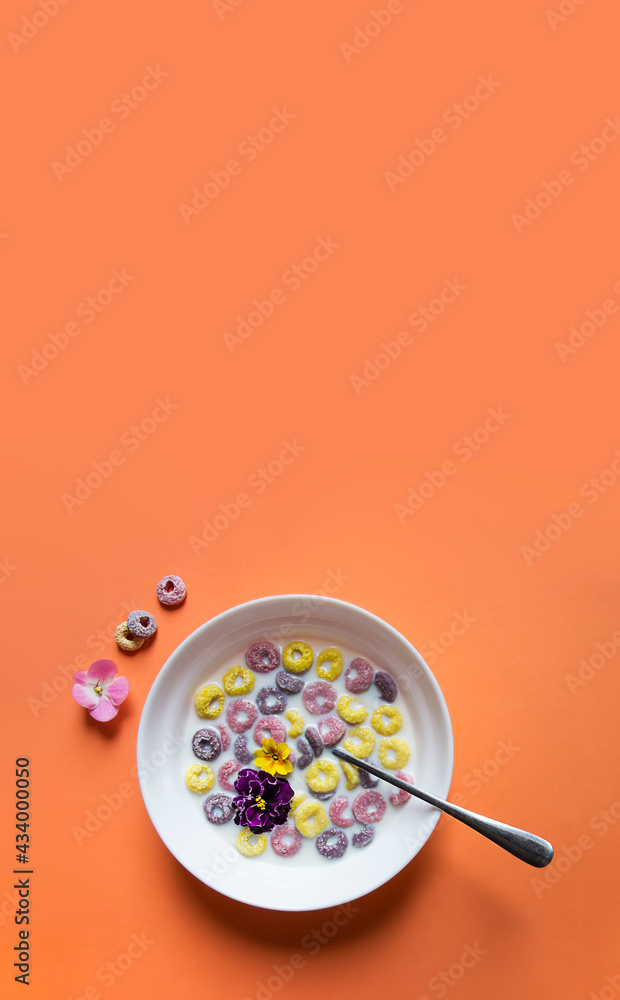 on a peach plain background a beautiful summer American breakfast: multi-colored flakes rings with milk and violet flowers of yellow pink and violet color. there is a spoon in the plate on a peach plain background a beautiful summer American breakfast: multi-colored flakes rings with milk and violet flowers of yellow pink and violet color. there is a spoon in the plate